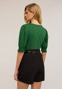 Top verde con mangas abullonadas y puños fruncidos, combinado con pantalones cortos negros de corte ajustado. Un cinturón negro elegante completa el atuendo casual.