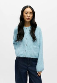 Svetlo modra pletena jakna z dolgimi rokavi, okroglim ovratnikom, srebrnimi gumbi in sprednjimi žepi. Mehak teksturiran material z puhastim zaključkom.