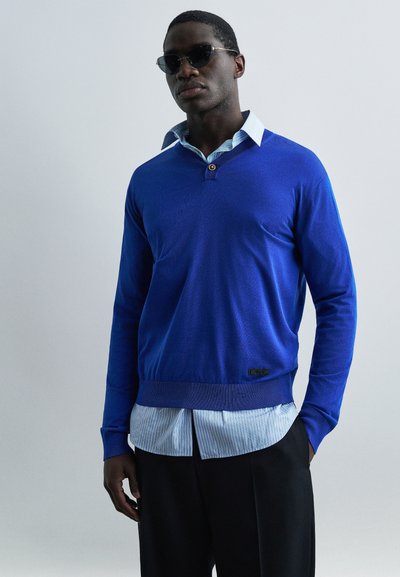 Homme portant des lunettes de soleil noires, un pull bleu par-dessus une chemise à col rayée bleue, et un pantalon noir, devant un fond uni gris clair.