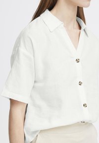 Camicia bianca di lino a maniche corte con colletto e sei bottoni tondi. Presenta una vestibilità rilassata e una texture sottile.