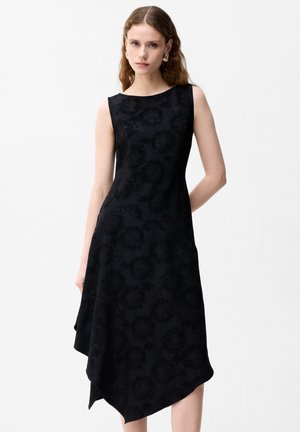 Młoda kobieta w czarnej, kwiecistej sukience jacquardowej bez rękawów z asymetrycznym dołem i złotymi kolczykami-kołami, stojąca na białym tle.