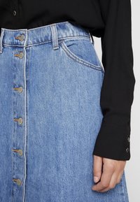 Jeanskjol i ljusblått med knäppning framtill. Har hög midja, sidofickor och kontrasterande sömmar. Svart långärmad skjorta syns.