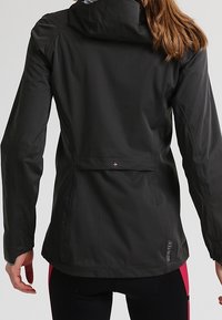 Mujer vistiendo una chaqueta de lluvia negra de Gore-Tex con capucha, ventilación trasera y mangas ajustadas, vista desde atrás.