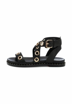 Sandalia negra con ojales dorados, ribete con tachuelas y una hebilla lateral. El diseño incluye dos correas frontales y una correa trasera en el tobillo para mayor soporte.