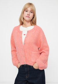 Przesadnie duży, miękki kardigan z dzianiny w jasnoróżowym kolorze, z okrągłym dekoltem, zapięciem na pięć guzików, dwoma przednimi kieszeniami i ściągaczami przy mankietach.