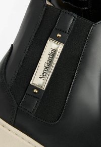 Bottine noire en cuir avec des panneaux élastiques sur les côtés, des accents en métal doré et une étiquette argentée bien visible. Finitions texturées et lisses.