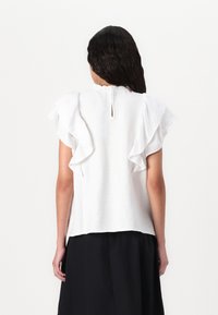 Blouse blanche avec des manches courtes froncées, un col rond avec un détail en forme de trou clé à l'arrière, faite en tissu léger texturé.