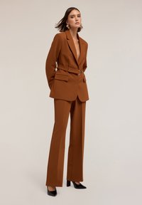 Bruine op maat gemaakte blazer en broekset met een riem met een ringgesp. Glad textuur. Zwarte hoge hakken complementeren de outfit.