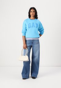 Sudadera azul con el logo "GAP", jeans de pierna ancha azules y un pequeño bolso blanco. La modelo está de pie contra un fondo liso.