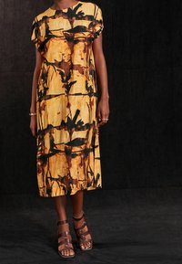 Vestido midi de estampado amarillo y negro con mangas cortas, aberturas laterales y cuello redondo. Sandalias de tiras marrones complementan el conjunto.