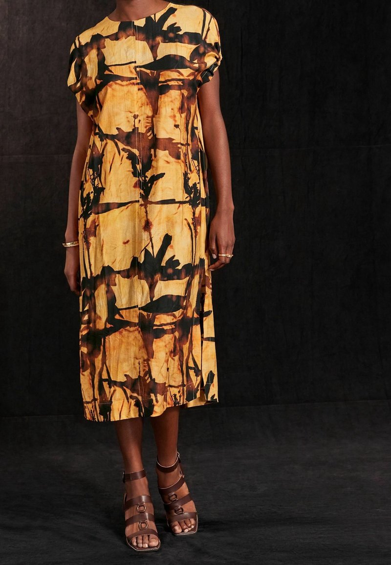 Vestido midi de estampado amarillo y negro con mangas cortas, aberturas laterales y cuello redondo. Sandalias de tiras marrones complementan el conjunto.