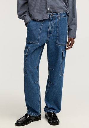 Blauwe denim cargobroek heeft een losse pasvorm, meerdere zakken en zichtbare stikdetails. Gecombineerd met zwarte loafers.