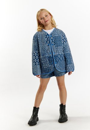 Blå quiltet jakke med geometriske mønstre, rund hals, kontrastfarvet blå trim, parret med denimshorts og sorte støvler.