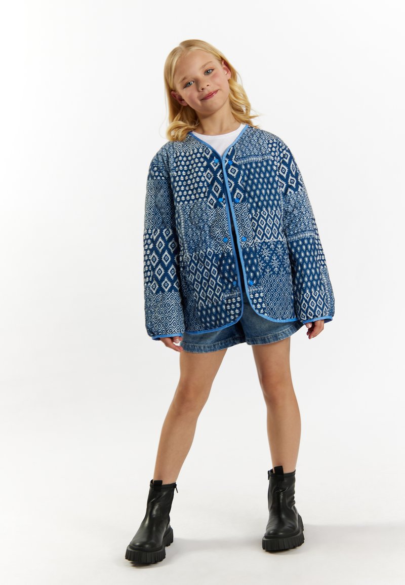 Blå quiltet jakke med geometriske mønstre, rund hals, kontrastfarvet blå trim, parret med denimshorts og sorte støvler.