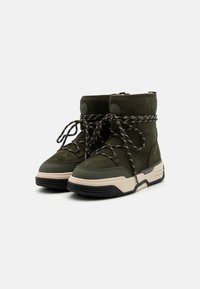 Bottines en daim vert avec des lacets à motif noir et blanc, une semelle en caoutchouc de couleur claire et des accents circulaires sur les côtés.