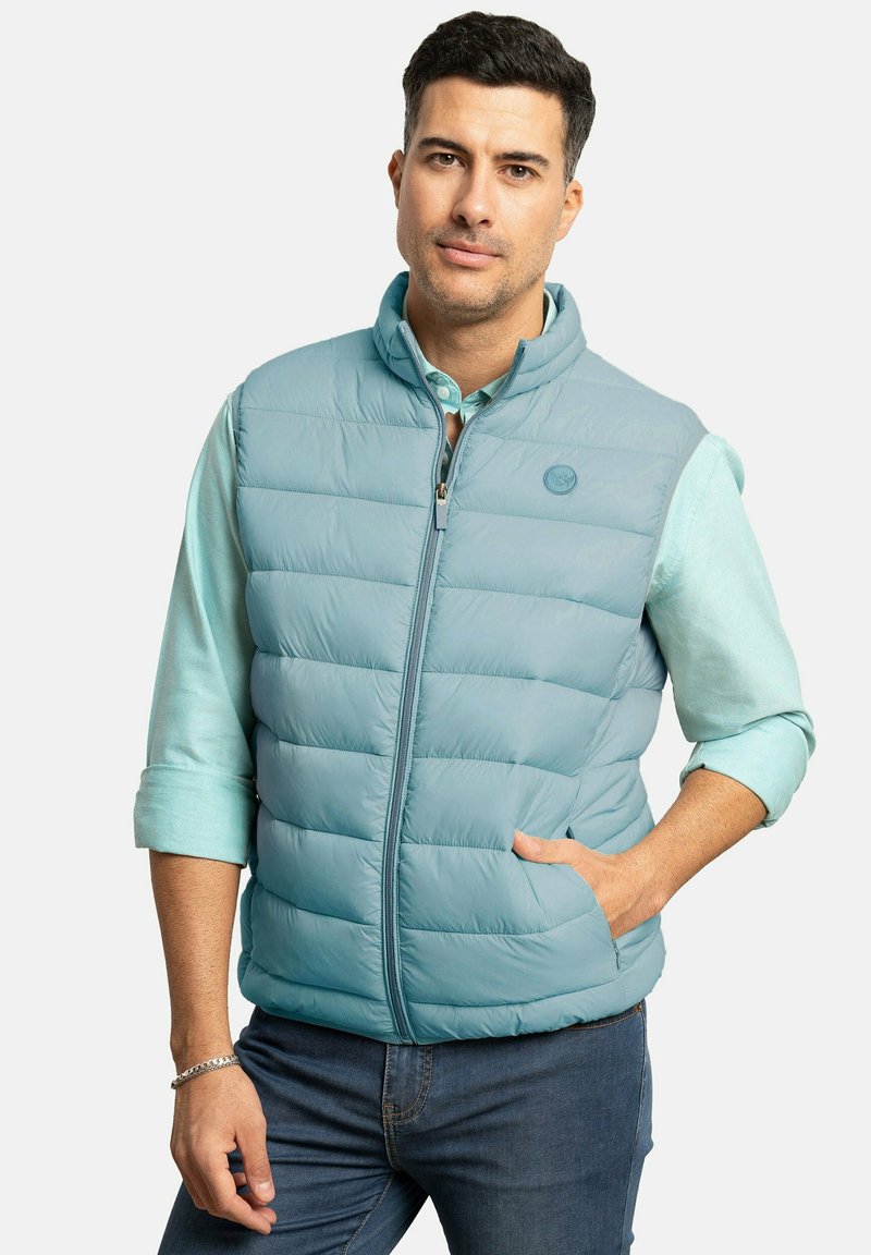 Chaleco acolchado azul claro con cuello alto y cremallera frontal, que presenta dos bolsillos laterales y un detalle de logo, usado sobre una camisa azul clara.
