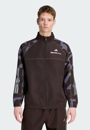 Veste de sport marron avec fermeture éclair, motif camouflage noir et gris, surpiqûres jaunes, et logo blanc sur la poitrine et les manches. Dotée de poignets élastiques.