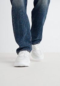 Hvite joggesko med tykk såle og teksturerte detaljer, brukt sammen med løstsittende blå denimjeans i en stående posisjon.