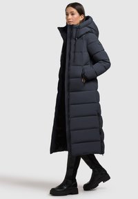 Cappotto imbottito lungo e di un blu navy scuro, con colletto alto e cappuccio, dotato di tasche laterali con zip e un motivo trapuntato. Indossato su pantaloni neri e stivali.