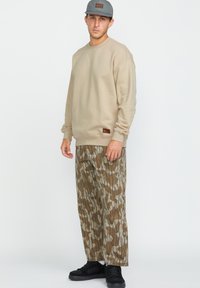 Sudadera de cuello redondo beige, pantalones camuflados en tonos de marrón y verde, zapatillas negras, gorra gris con parche de logo.