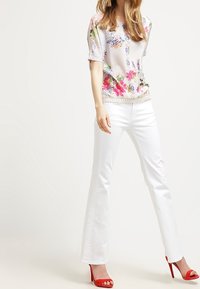 Floral-print short-sleeve blouse in light fabric, paired with white flared trousers. Model wears red high-heeled sandals.