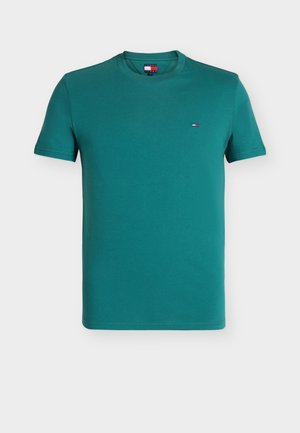 Mand stående iført en turkis kortærmet skjorte, lyseblå jeans, hvide sneakers, der holder en sammenfoldet lysegrå yogamåtte i højre hånd.