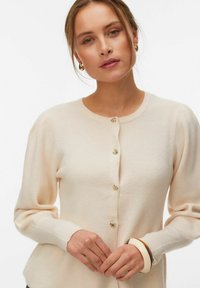 Femme portant un cardigan crème avec des boutons dorés, des boucles d'oreilles créoles dorées et des bracelets crème et marron, posant devant un fond blanc uni.