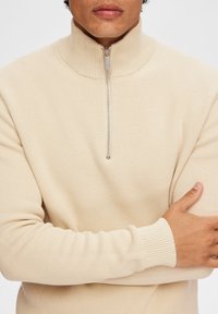 Beiger, gerippter Strickpullover mit hohem Kragen und halbem Reißverschluss. Verfügt über schmale Bündchen und eine strukturierte Oberfläche, geeignet für Freizeitkleidung.