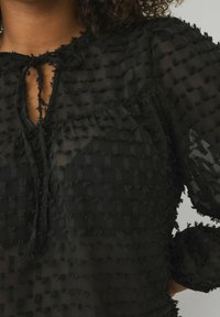 Zwarte doorzichtige blouse met een textuur en geweven patroon met franjes. Rond de hals met strikdetail en lange, opgeblazen mouwen.