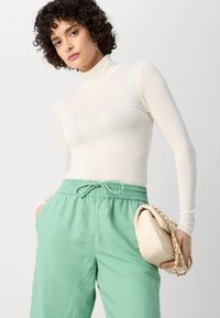 White long-sleeve turtleneck top paired with light green drawstring pants. Holding a cream-colored handbag with chain strap.