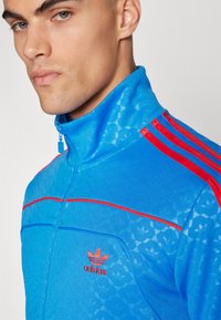 Zila zip-up jaka ar sarkanām svītrām un Adidas logo, ar teksturētu rakstu un augstu apkakli.