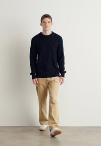 Maglione lavorato a maglia blu navy con un motivo a diamante, abbinato a pantaloni beige e sneakers bianche, indossato da una persona in piedi su una superficie neutra.