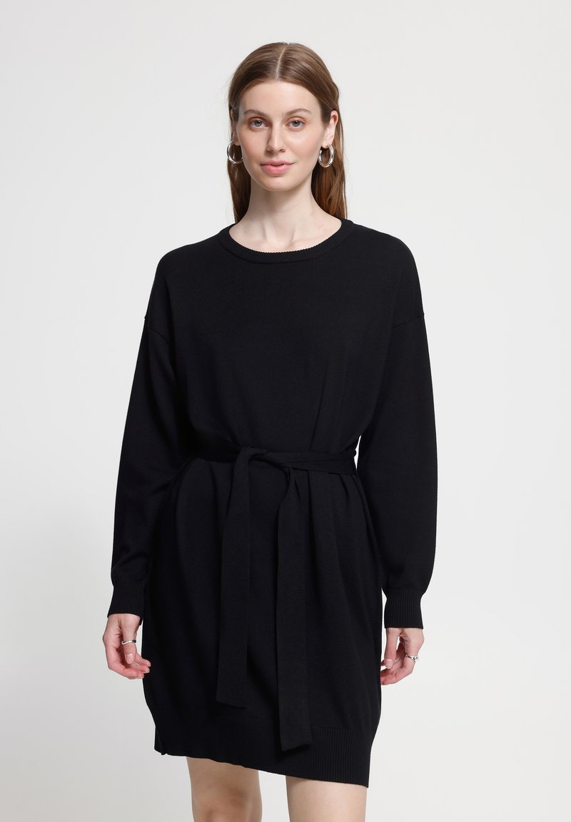 Jeune femme aux longs cheveux bruns portant une robe noire à manches longues ceinturée et des boucles d'oreilles argentées en anneaux, debout devant un fond clair uni.