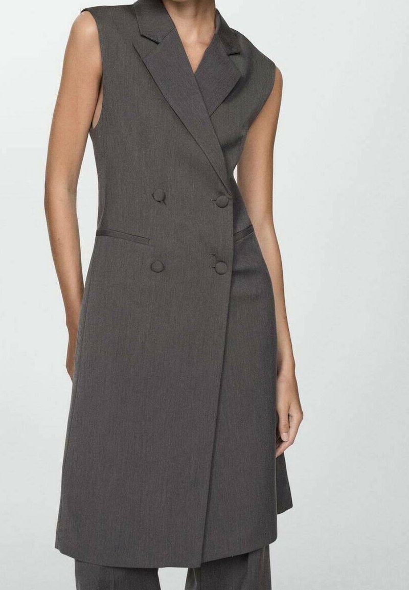 Robe blazer sans manches gris foncé à double boutonnage avec revers et fente latérale, portée avec un pantalon assorti sur une personne debout.