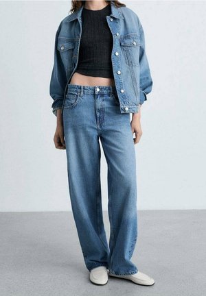 Person iført en lyseblå denimjakke og vide jeans kombineret med en sort cropped top og hvide slip-on sko, står på en grå gulv.