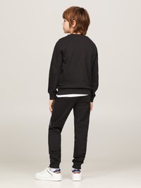 Schwarzer Sweatshirt und passende Jogginghose aus weichem Material, mit gerippten Bündchen und einem Logodetail am Bein. Weiße Sneakers vervollständigen das Outfit.