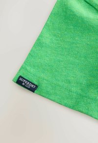 Coin en tissu vert d'un vêtement avec des coutures visibles et une étiquette de marque noire "SUPERDRY & CO" cousue près de l'ourlet.