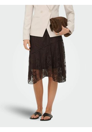 Žena v béžovom blazeri, čiernej čipkovanej midi sukni, čiernych šľapkách drží zloženú hnedú semišovú kabelku.