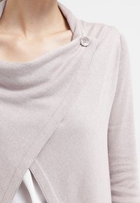 Lichtroze cardigan met een gedrapeerde halslijn, voorzien van een enkele doorzichtige knoop en een wikkelontwerp. Zachte, gestructureerde stof, lange mouwen.