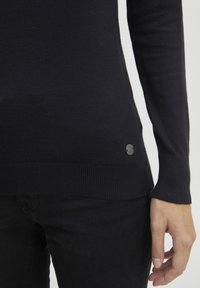 Schwarzer, gestrickter Pullover mit langen Ärmeln, gerippten Bündchen und Saum, der ein kleines metallisches Logo-Detail an der unteren Seite aufweist.