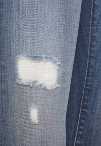 Tessuto di denim azzurro chiaro con due toppe rettangolari strappate che mostrano i fili bianchi sottostanti.