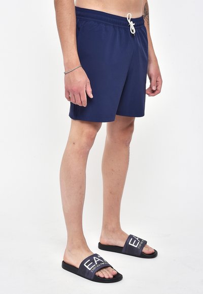 Costume da bagno blu navy con elastico in vita e cordino. Abbinati a ciabatte con logo bianco su sfondo scuro.