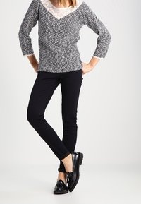 Svartvit mönstrad topp med spetskantad halsringning, tillsammans med svarta trånga byxor och blanka svarta loafers.