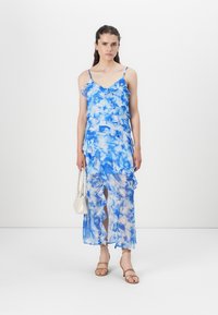 Μπλε φόρεμα με σχέδιο, φτιαγμένο από ριχτά στρώματα, με τιράντες spaghetti και σκίσιμο. Συνδυάζεται με μια λευκή τσάντα και γυμνές σανδάλες. Απαλή και ελαφριά υφή.