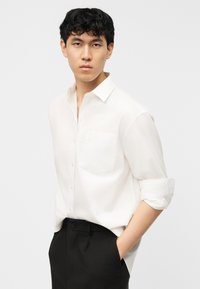 Νεαρός άντρας με κοντά μαύρα μαλλιά, ντυμένος με λευκό πουκάμισο με κουμπιά και μουσελίνες στα μανίκια και μαύρο παντελόνι, στέκεται με τα χέρια στις τσέπες.