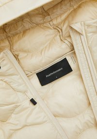 Beige gewatteerde jas met een gladde textuur, voorzien van een centrale rits, verstelbare capuchon en een label met de tekst "PeakPerformance," maat S.