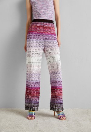 Strikkede bukser med vide ben og et zigzagmønster i nuancer af rød, lilla og hvid, med en sort taljebånd og flerfarvede stropper på skoene.