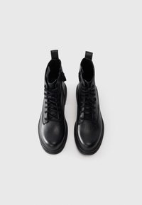 Par de botas de tobillo de cuero negro con cordones, tiradores y cremalleras laterales sobre un fondo blanco liso.