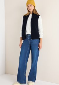Marineblauer Strickwesten, weiß gepunkteter Pullover, hellblaue weit geschnittene Jeans und eine senfgelbe Mütze, kombiniert mit beigen Sneakers.