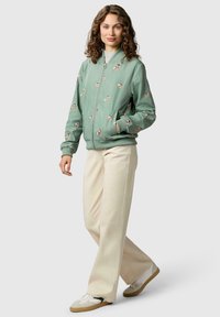 Kobieta o kręconych włosach, ubrana w zieloną, kwiatową kurtkę bomberkę, beżowe spodnie o szerokich nogawkach i białe trampki, stojąca na tle jednolitego tła.